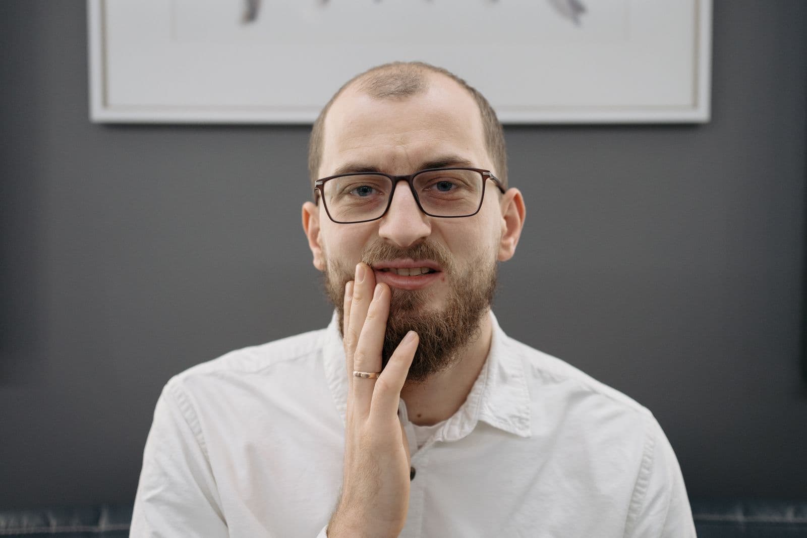 A man with eyeglasses and a beard shows discomfort from a toothache indoors.