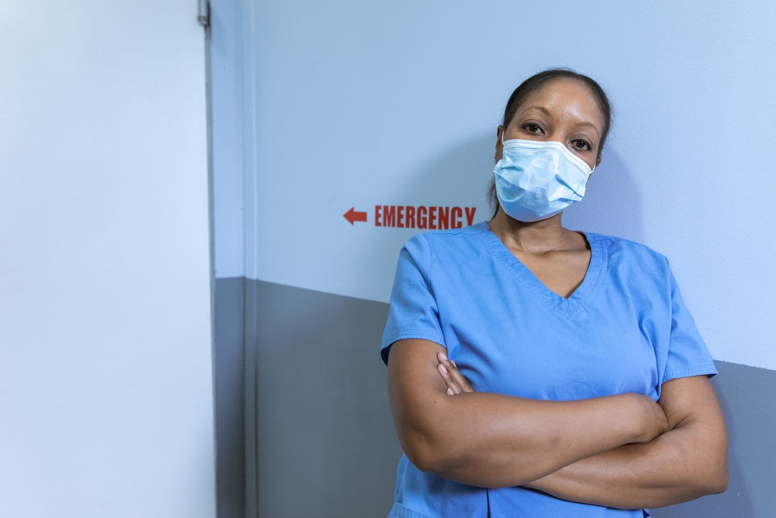 A nurse in scrubs stands in a hospital corridor under an emergency sign.